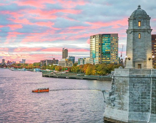 A scenic view of a river at sunset, with a boat, city skyline, and colorful clouds reflecting in the water.
