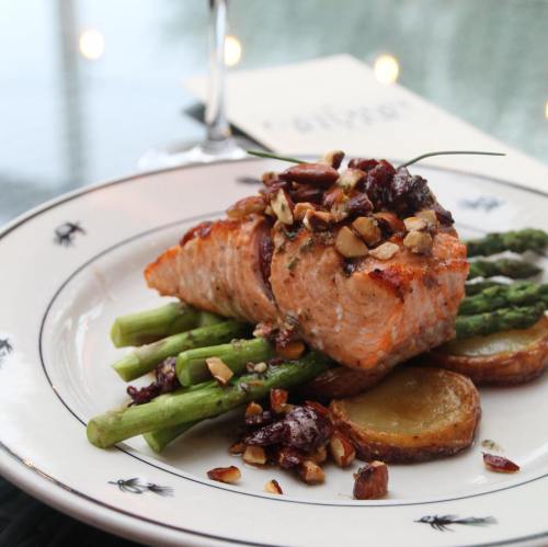 A plated dish featuring salmon on asparagus and potatoes, topped with nuts and herbs, with a glass in the background.