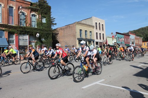 A large group of cyclists in colorful jerseys riding down a street lined with historic buildings on a sunny day.