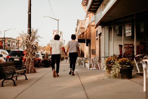 Two people walk hand in hand down a sidewalk, surrounded by shops and autumn decorations.