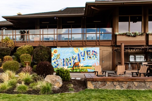 A colorful mural reading "Greetings From Sunriver, Oregon" on a building surrounded by greenery and outdoor seating.