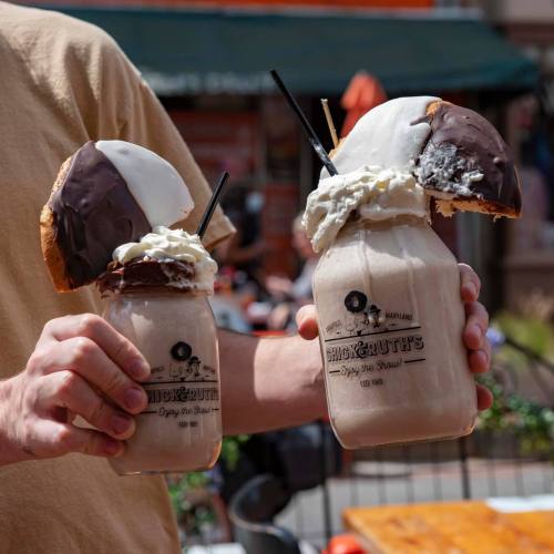 Two large milkshakes topped with whipped cream and cookies, held by a person outdoors.