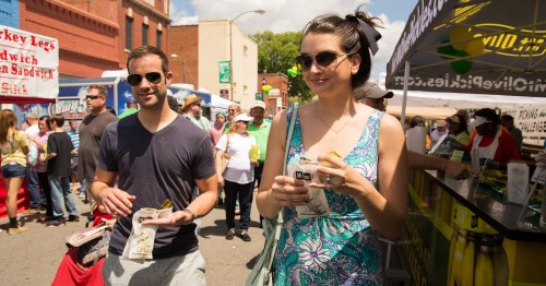 A couple walks through a busy street fair, holding food and drinks, surrounded by vendors and a lively crowd.