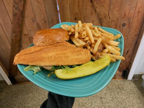A plate with a breaded fish fillet, a bun, fries, and a pickle, held against a rustic wooden background.
