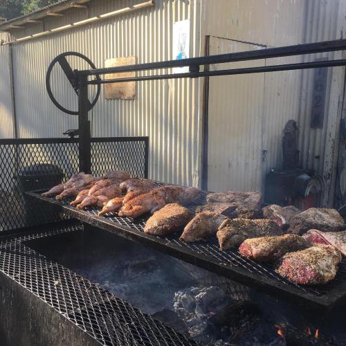 Grilled meats on a large barbecue grill, with smoke rising against a rustic metal backdrop.