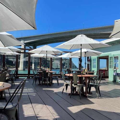 Outdoor dining area with tables and umbrellas, overlooking water and a bridge under a clear blue sky.