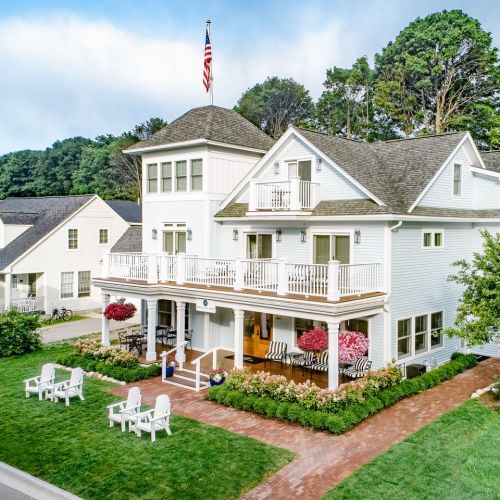A charming white house with a porch, surrounded by green trees and a well-maintained lawn, featuring outdoor seating.