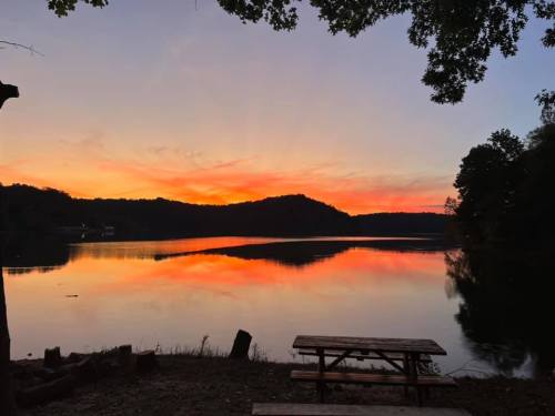 A serene lake at sunset, reflecting vibrant orange and pink hues, with a picnic table in the foreground and trees along the shore.