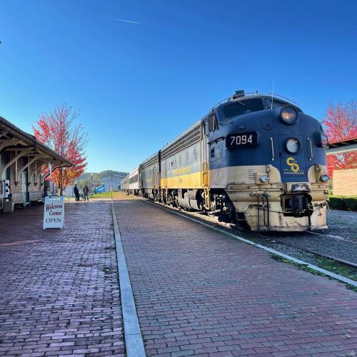 A vintage train at a station with colorful autumn trees and a sign for the open welcome center.