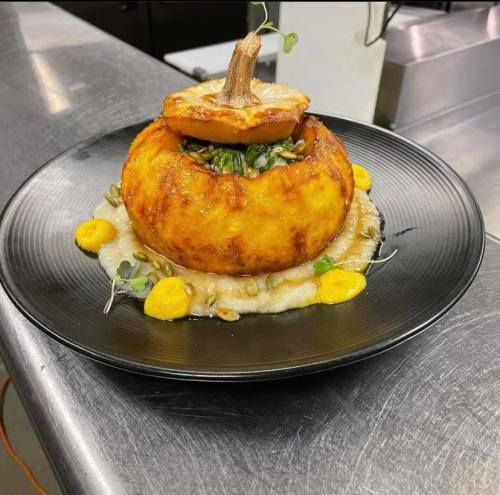 A beautifully plated dish featuring a round, baked pumpkin filled with greens, served on a bed of creamy puree and garnished.