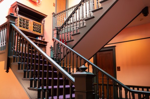A vintage staircase with dark wood railings and purple carpet, leading to upper floors in a warmly lit hallway.