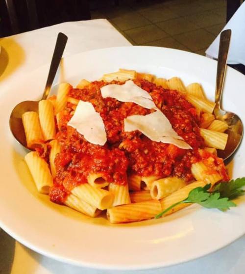 A plate of rigatoni pasta topped with meat sauce and cheese, garnished with fresh herbs, served with two spoons.