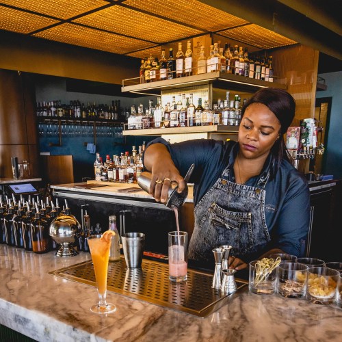 A bartender in an apron pours a cocktail at a stylish bar with shelves of liquor in the background.