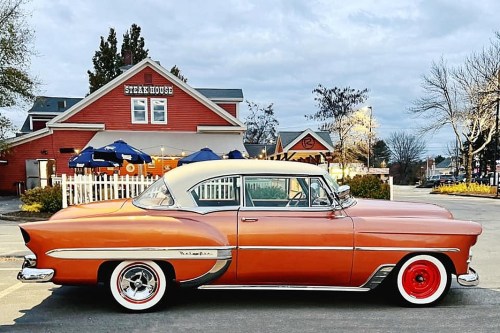 A vintage orange and cream car parked in front of a steakhouse, with a cloudy sky in the background.