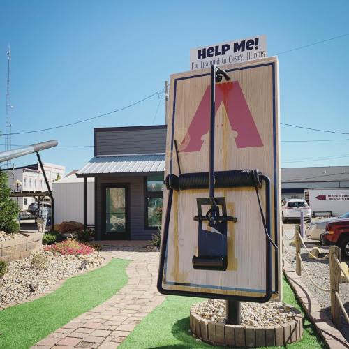 A large sign with the text "Help Me!" and a trap, set in a small town with a green pathway and buildings in the background.