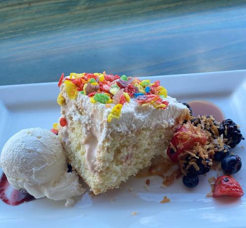 A slice of cake topped with colorful cereal, served with a scoop of ice cream and mixed berries on a white plate.