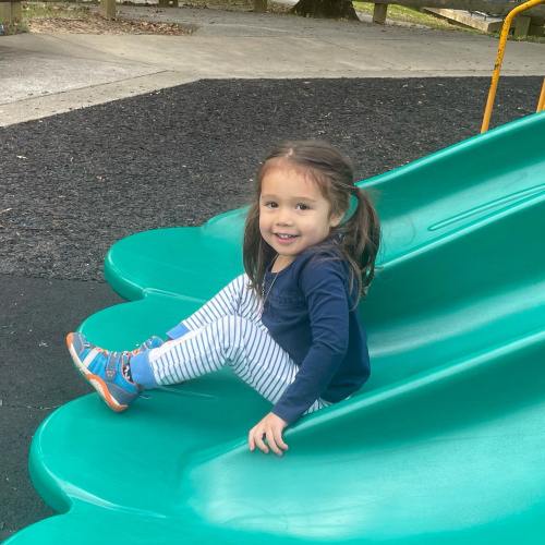 A young girl sits on a green slide at a playground, smiling and wearing a blue sweater and striped pants.