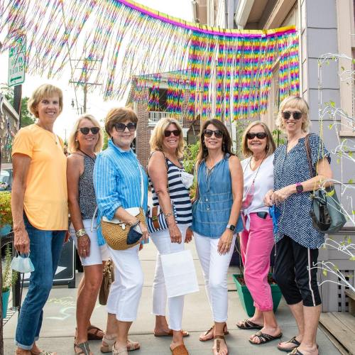 A group of seven women poses together outdoors, smiling in front of colorful decorations.