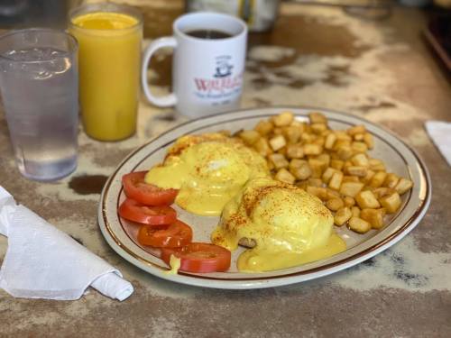 A plate of eggs Benedict with tomatoes and hash browns, accompanied by a glass of water, orange juice, and coffee.