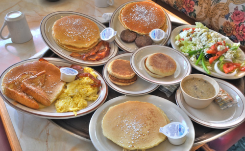 A tray filled with various breakfast items: pancakes, eggs, sausage, bacon, a salad, and a cup of soup.
