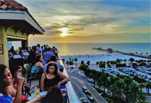 A vibrant sunset over the ocean, viewed from a rooftop bar with people enjoying drinks and the beach below.