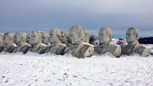 Row of large stone busts partially covered in snow, set against a cloudy sky.
