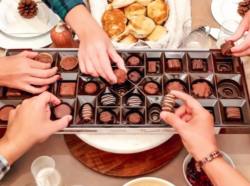 A group of hands reaching for assorted chocolates in a box, with baked goods and a festive table setting in the background.