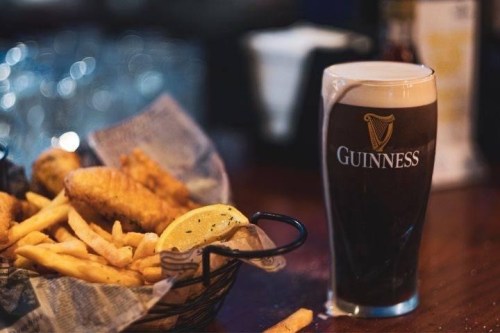 A pint of Guinness next to a basket of fish and chips with lemon wedges on a wooden table.