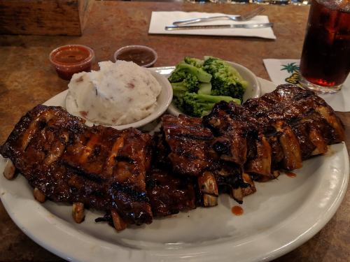 A plate of barbecue ribs with mashed potatoes, broccoli, and dipping sauces, served in a casual dining setting.