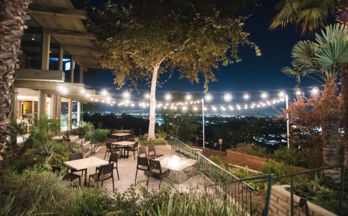 Outdoor dining area with string lights, surrounded by greenery and a view of the city at night.