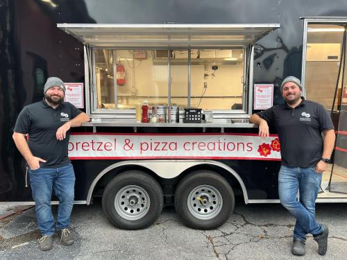Two men stand in front of a food truck labeled "Pretzel & Pizza Creations," smiling and posing for the photo.
