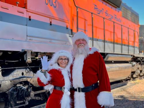 A man and woman in Santa costumes pose cheerfully in front of a bright orange train.