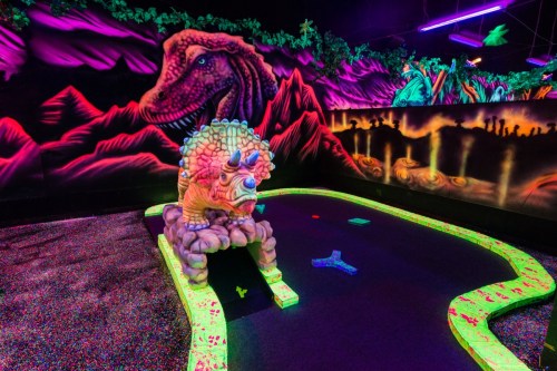 A vibrant glow-in-the-dark mini-golf course featuring a dinosaur-themed sculpture and colorful neon decorations.