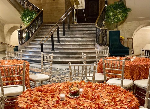 Elegant venue with tables adorned in orange leaves, surrounded by greenery and a grand staircase in the background.