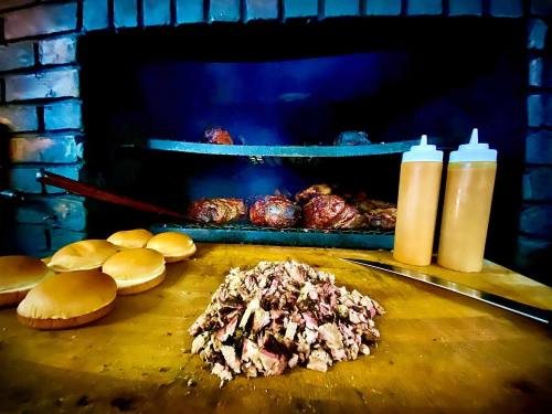 A wooden table with chopped meat, buns, and two squeeze bottles in front of a smoky barbecue grill.