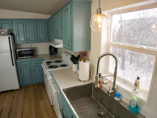 A bright kitchen with teal cabinets, a stainless steel sink, and a window overlooking a snowy landscape.