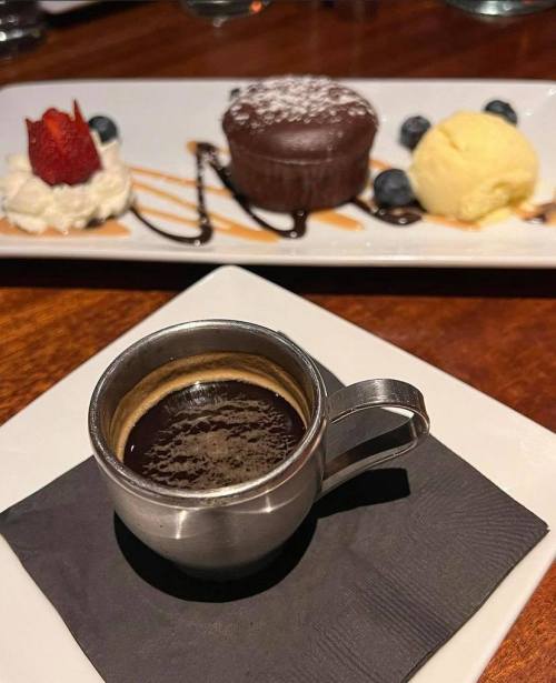 A small metal cup of espresso on a black napkin, with a dessert plate featuring chocolate cake, ice cream, and berries in the background.