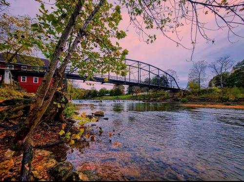 A serene river scene with a bridge, trees, and a colorful sky at sunset, reflecting on the water's surface.