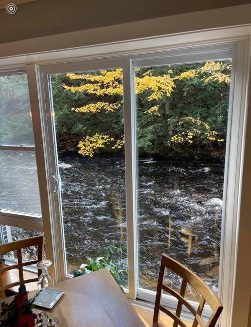View of a river through large windows, framed by autumn leaves and a cozy dining area with wooden chairs.