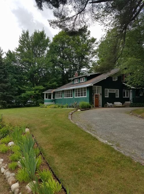 A green house surrounded by trees and a well-maintained lawn, with a gravel path leading to the entrance.