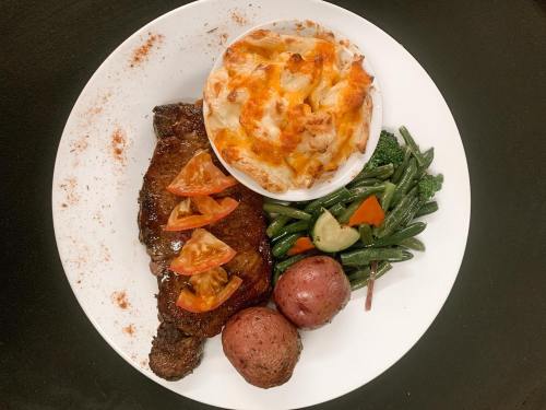 A plate featuring grilled steak with sliced tomatoes, creamy pasta, red potatoes, and mixed vegetables.