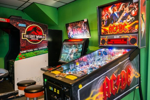 A colorful arcade room featuring an AC/DC pinball machine and a Jurassic Park arcade game against a green wall.