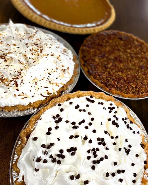 Three delicious pies: one with chocolate chips, one with pecans, and a pumpkin pie in the background. All topped with whipped cream.