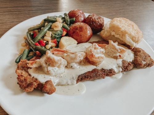 A plate featuring fried fish topped with shrimp and creamy sauce, alongside roasted potatoes, mixed vegetables, and a biscuit.