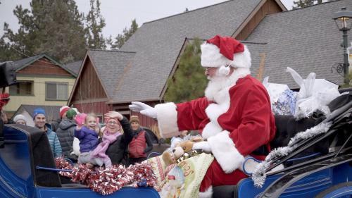 Santa Claus waves from a decorated sleigh, surrounded by excited children and a festive crowd in a winter setting.