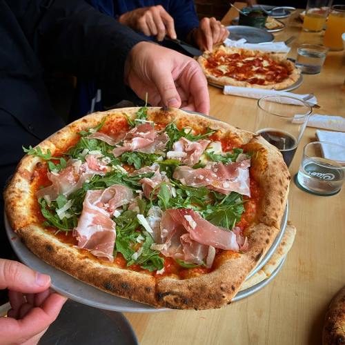 A person holds a pizza topped with arugula and prosciutto, with other pizzas and drinks visible on the table.