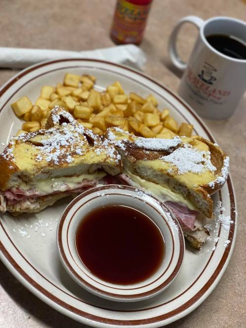 A plate with a stuffed sandwich, powdered sugar, a side of cubed potatoes, and a small bowl of syrup. Coffee in the background.