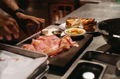 A chef's hand arranges sliced meats on a wooden board, with toast and condiments in the background.