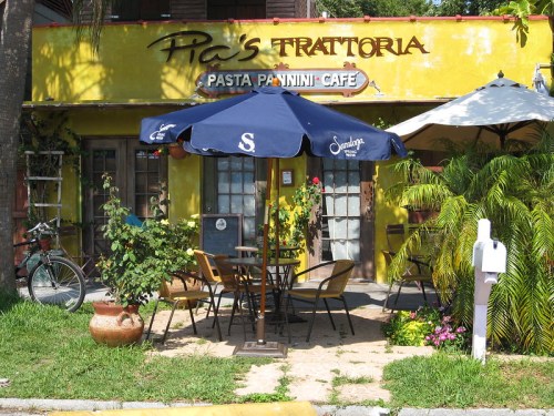 A cozy outdoor dining area with tables, umbrellas, and lush greenery in front of a yellow restaurant.
