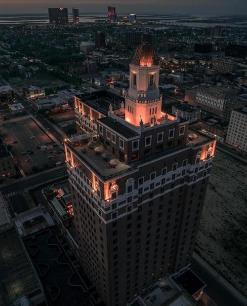 Aerial view of a tall, illuminated building at dusk, surrounded by a cityscape and distant water.